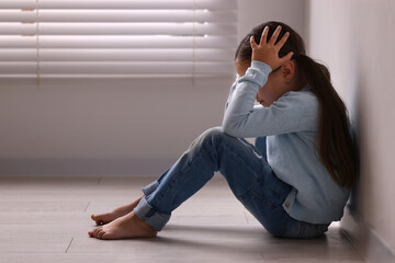 Child abuse. Upset little girl sitting on floor near light wall indoors
