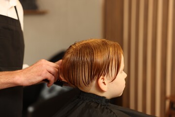 Naklejka premium Professional hairdresser combing boy's hair in beauty salon, closeup