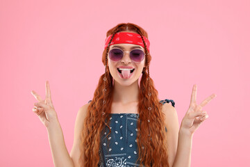 Stylish young hippie woman in sunglasses showing V-sign on pink background