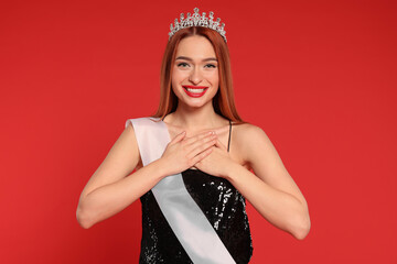 Beautiful young woman with tiara and ribbon in dress on red background. Beauty contest