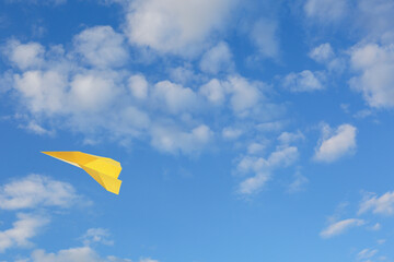 Yellow paper plane flying in blue sky with clouds