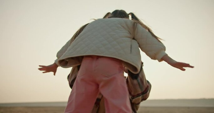 A happy middle-aged brunette man with gray hair in a plaid shirt lifts his little daughter a blonde girl in a white jacket and spins with her during his vacation on a hike on a deserted seashore in a