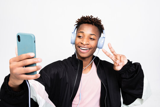 Cheerful attractive dark skinned guy hipster student man standing over white background in studio isolated smiling holding smartphone cell phone in hands taking picture while having shooting process.