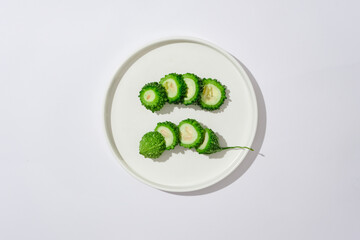 On a white background and a round ceramic dish container fresh slices of bitter melon. Advertising scene. Bitter gourd is known as a simple and effective natural way to beautify the skin