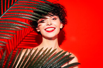 Fashion beauty portrait of young smiling brunette woman with evening stylish  makeup and perfect clean skin. Sexy model with curly short hair posing in studio. With red bright lips. Tropical palm leaf