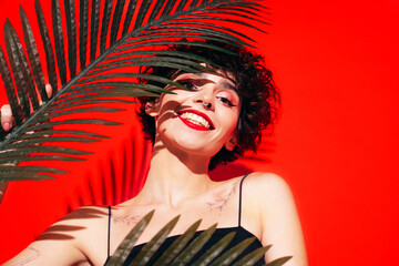 Fashion beauty portrait of young smiling brunette woman with evening stylish  makeup and perfect clean skin. Sexy model with curly short hair posing in studio. With red bright lips. Tropical palm leaf