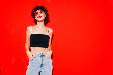 Young beautiful smiling female in trendy summer black tank top. Carefree woman posing near red wall in studio with curly hairstyle. Positive model having fun, going crazy. In sunglasses
