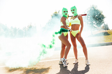 Two beautiful sexy women in green underwear. Models wearing bandit balaclava mask. Hot seductive female in nice lingerie posing in the street at sunny day. Crime and violence. Hold green smoke bomb - obrazy, fototapety, plakaty