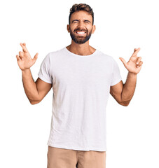 Young hispanic man wearing casual white tshirt gesturing finger crossed smiling with hope and eyes closed. luck and superstitious concept.