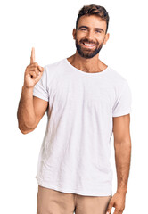 Young hispanic man wearing casual white tshirt showing and pointing up with finger number one while smiling confident and happy.