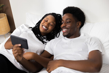 Delighted happy american black family sit on bed with cell phone smiling look at smartphoone screen...