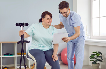 Patient with injury undergoes medical treatment and physiotherapy rehabilitation workouts to regain mobility of joints. Happy fat woman with injured leg looks at clipboard that man specialist shows