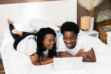 Young african american black couple lying on bed in front of laptop watching series movie tv show.