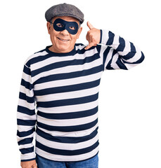 Senior handsome man wearing burglar mask and t-shirt smiling doing phone gesture with hand and fingers like talking on the telephone. communicating concepts.