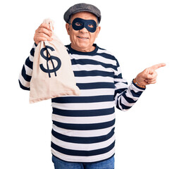 Senior handsome man wearing burglar mask holding money bag smiling happy pointing with hand and...