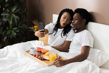 Happy black couple lying in bed having breakfast beautiful African American wife taking selfie...