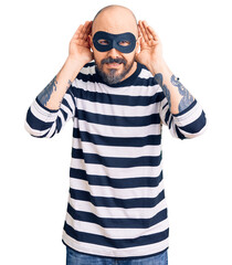 Young handsome man wearing burglar mask trying to hear both hands on ear gesture, curious for...
