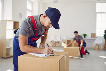 Worker from professional man and van delivery company writes something on paper while removing stuff on house moving day. Loader in uniform workwear and baseball cap fills out invoice on cardboard box - obrazy, fototapety, plakaty