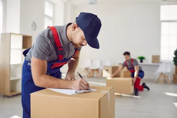 Fotobehang Worker from professional man and van delivery company writes something on paper while removing stuff on house moving day. Loader in uniform workwear and baseball cap fills out invoice on cardboard box © Studio Romantic