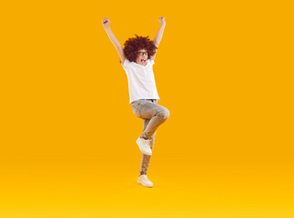 Fototapeta na wymiar Preteen smiling boy happily jumping with his hands raised. Excited boy in curly red wig wearing white t-shirt, jeans and sneakers having fun over yellow studio background. Good mood, joy, success