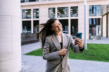 Fototapeta premium Cheerful young woman in business suit makes video call by phone walking by street toothy smiles happy goes home after work. Female employee happy outside.