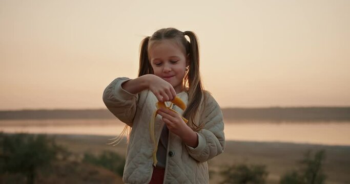 Portrait of a little blonde girl with blue eyes in a white jacket who peels a banana and eats it during her picnic outside the city in the evening in summer