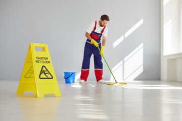 Caution wet floor sign with male young cleaner mopping empty room or office in background. Man janitor with mop in hands and bucket from cleaning service washing gray floor in uniform. - obrazy, fototapety, plakaty