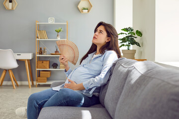 What a terrible day. Too hot at home. Young pregnant woman, tired of extreme summer heatwave, sitting on sofa in overheated living room, dreaming of fresh breeze, and refreshing herself with hand fan