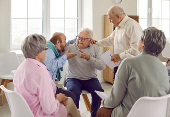 Two old men argue, exchange negative opinions, offend, insult each other while sitting with other senior people at talking therapy session devoted to constructive communication and conflict resolution