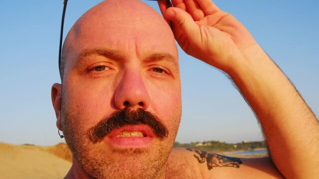 caucasian male bald alopecia with moustache wearing sunglasses at the beach during sunset