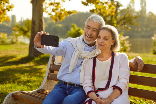 Happy old senior couple enjoying good sunny summer day relaxing on bench in beautiful park garden with green trees in background, using modern smart cell phone to take selfie with romantic life moment