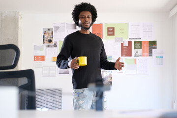 Afro man with casual style in his office talking and gesturing during a video call while holding a cup of coffee. In the background is the moodboard with projects in progress of the business. In the f