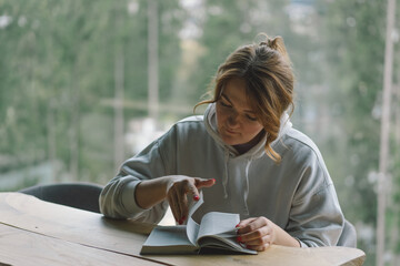 A Woman reads the book and drinks coffee from a thermos cup near the beautiful panoramic window against the background of the forest.