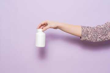 White plastic bottle in female hand in lilac background. Packaging for pills, capsules or supplements.