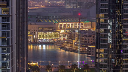 Aerial view of Dubai Fountain in downtown with palms in park next to shopping mall and souq day to night timelapse, UAE