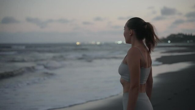 A sporty girl runs to the beach at the end of her workout and looks at the sunset