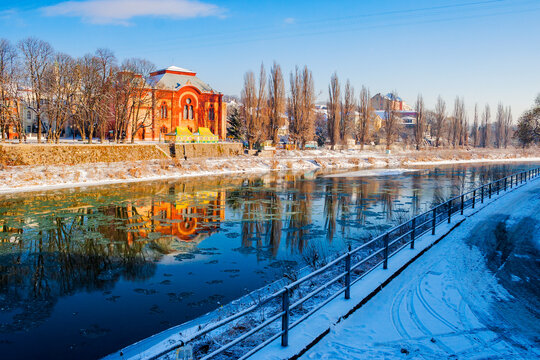 uzhgorod cityscape in winter. urban landscape with snow covered embankment. red building of a former synagogue reflecting in the river. cold weather on a sunny day