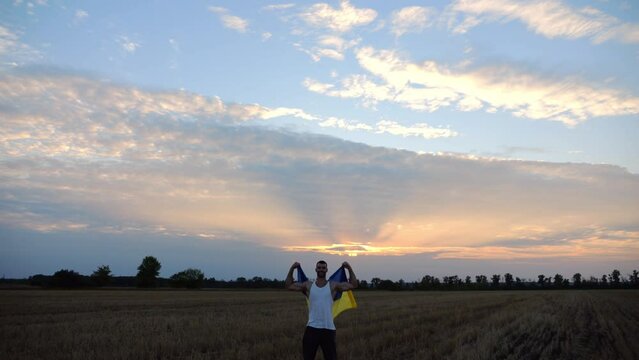 Ukrainian sportsman stands on barley meadow showing biceps with blue-yellow banner in hands. Young guy raised flag of Ukraine above head on wheat field at sunset. Victory against russian aggression