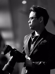 Vertical black and white portrait of an Asian man with short, cool black hair, wearing glasses, wearing a black suit, business man, holding a camera. with a background of light and shadow