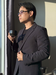 Naklejka premium Vertical portrait of an Asian man with short, cool black hair, wearing glasses, wearing a black suit and businessman, drinking coffee with a shadow of a window in the background.
