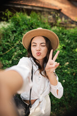 Portrait of young lady wear summer hat smiling showing v-sign taking selfie outdoors urban city park
