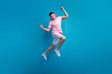 Fototapeta premium Full size portrait of overjoyed active man jumping raise fists listen music headphones isolated on blue color background