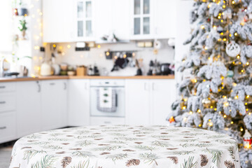 Round empty table to demonstrate the product with with tablecloth with pine needles and cones print, space for text in white kitchen, modern interior with a Christmas tree. New Year, Christmas