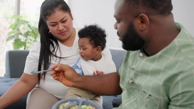 Asian mother feeding her 9 months old her cute little baby and African American helping for holding food plate At Home. Photo series of family, kids and happy people concept. Parents feed kids.