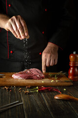 The cook sprinkles salt into meat steak for cooking for barbecue lunch. Working environment on the kitchen table with fragrant rosemary and garlic.