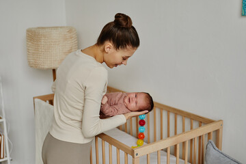 Side view at loving young Caucasian mom putting her happy newborn to cot for sleep, copy space
