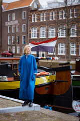 Obraz premium A woman in a stylish blue coat on a canal with ships and a waving flag of France. Feelings of passion for travel and adventure. Residential area in the Netherlands.
