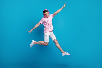 Full size photo of cheerful overjoyed man jump arms wings flying listen music headphones isolated on blue color background