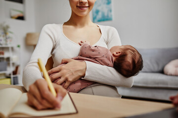 Cropped shot of napping baby in arms of smiling mother writing in notebook, copy space