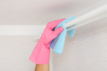 Woman hand in pink rubber protective glove holding blue dry rag and wiping white curtain rods at...